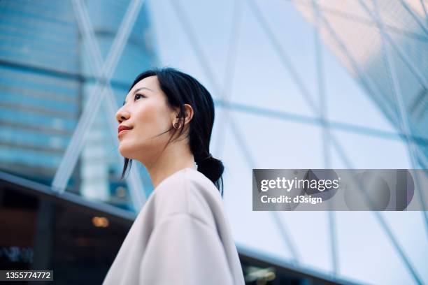 confident and ambitious young asian businesswoman looking away, standing in front of contemporary corporate skyscrapers in financial district in the city. girl power. career plan. woman at work. female leadership concept - looking up ストックフォトと画像