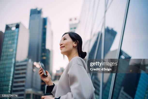 confident and ambitious young asian businesswoman with smartphone looking away, standing in front of contemporary corporate skyscrapers in financial district in the city. lifestyle and technology. woman at work. female leadership concept - will call stock pictures, royalty-free photos & images