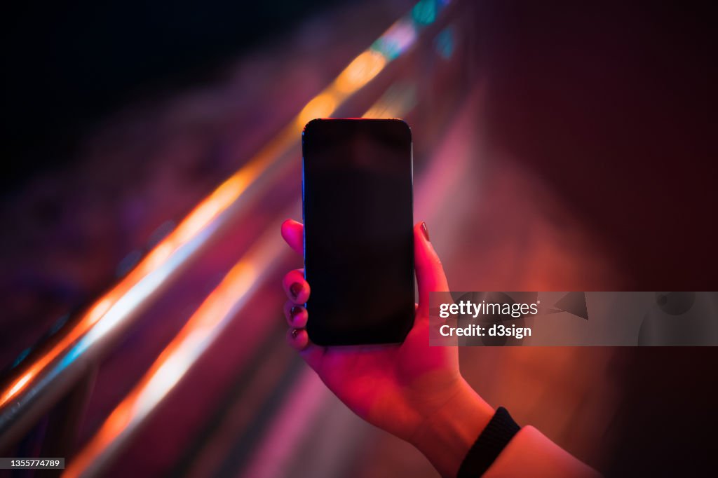 A woman lit by red neon coloured lights and close up of her hand using smartphone in downtown prosperous city street at night. Smartphone with blank screen for design mockup