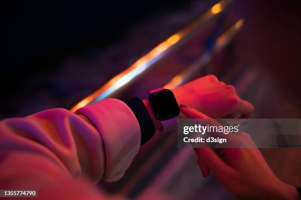 a woman lit by red neon coloured lights and over the shoulder view of her hand using smartwatch in downtown prosperous city street at night. smartwatch with blank screen for design mockup - reloj inteligente fotografías e imágenes de stock