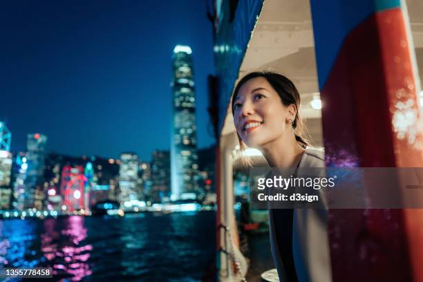 beautiful smiling young asian woman looking out through the window, enjoying spectacular illuminated night view of the city while travelling by ferry - entertainment occupation stockfoto's en -beelden