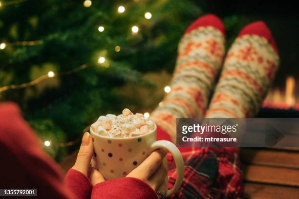 image of unrecognisable woman wearing red tartan pyjamas and christmas themed patterned socks, feet on coffee table, holding spotty, mug of hot chocolate, topped with whipped cream and mini marshmallows, row of gas fire flames in background - feet up stock pictures, royalty-free photos & images
