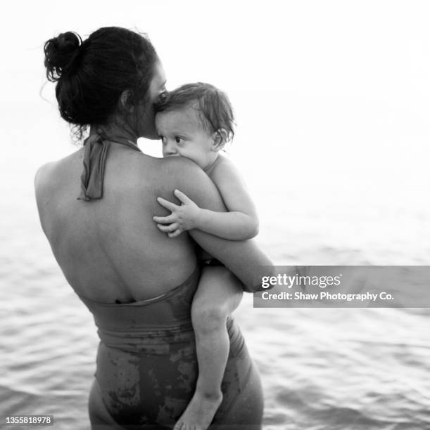 black and white photo of a mother holding her toddler son in the water. they are both in bathing suits and the mother's back is to us with a bun in her hair. the sun is peeking over her shoulder and cuddled in.  the horizon is over the water. - monochrome fotografías e imágenes de stock