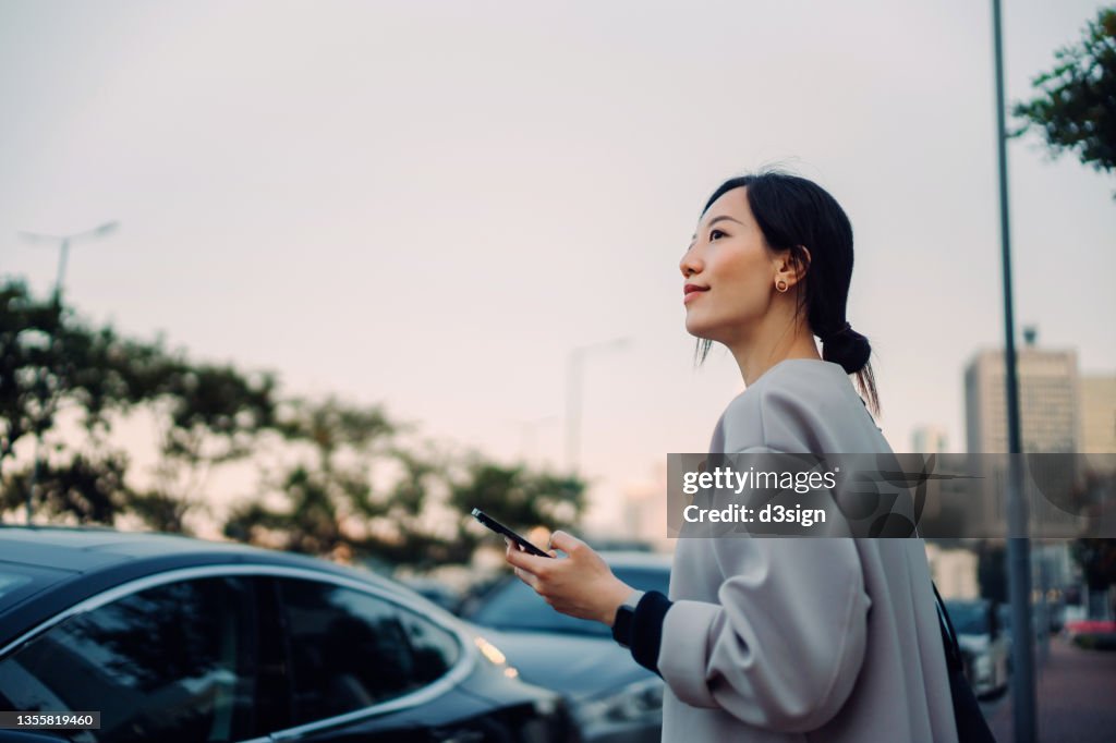 Confident and professional young Asian businesswoman walking to her car, holding smartphone on hand. Looking away against urban cityscape. Business on the go concept