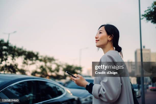 confident and professional young asian businesswoman walking to her car, holding smartphone on hand. looking away against urban cityscape. business on the go concept - people in car stockfoto's en -beelden