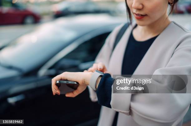 cropped shot of young asian businesswoman holding smartphone, checking time on smartwatch, standing next to her car in car park in the city. business on the go concept - rush shopping stock pictures, royalty-free photos & images