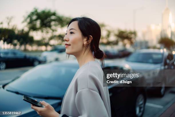 confident and professional young asian businesswoman walking out of her car, holding smartphone on hand. looking away against urban cityscape. business on the go concept - management car smartphone stock pictures, royalty-free photos & images