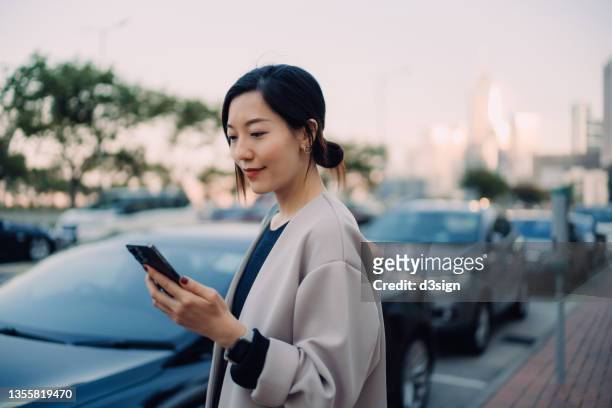 confident and professional young asian businesswoman using smartphone while walking to her car in the city, with urban cityscape in background. business on the go concept - market intelligence imagens e fotografias de stock
