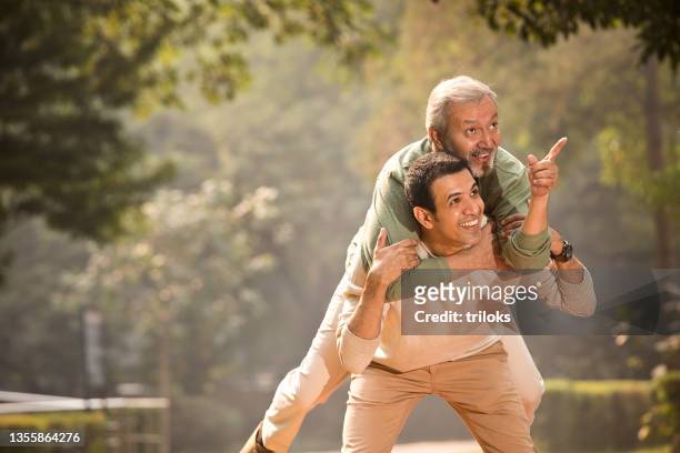mid adult man piggybacking his old father at park - piggyback stock pictures, royalty-free photos & images