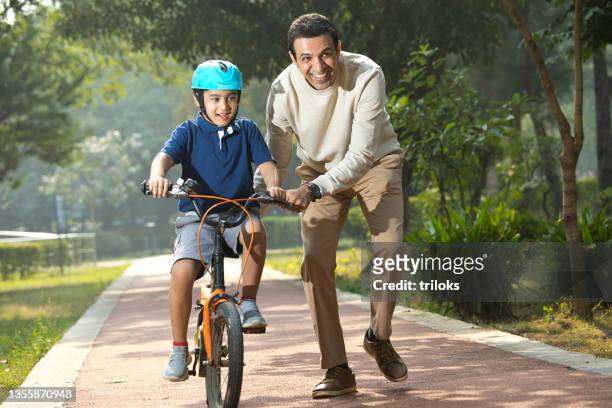 boy learning bicycle with assistance of father at park - sports india stockfoto's en -beelden