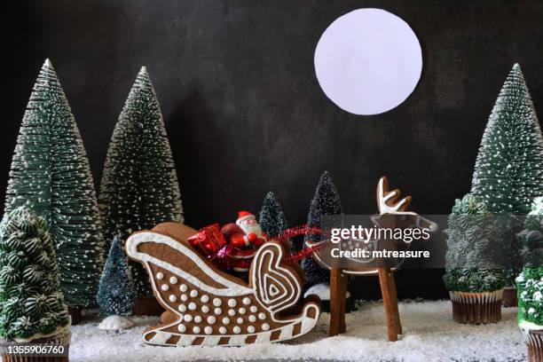 image of homemade, gingerbread cookie reindeer and sleigh, decorated with white royal icing, containing father christmas with gifts surrounded by night-time christmas forest scene of model fir and cupcake christmas trees, black background, full moon - cookie monster stockfoto's en -beelden