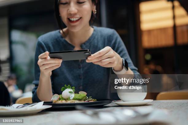 joyful young asian woman taking photos of delicious food freshly served on dining table with smartphone and sharing to social media in a sidewalk cafe. outdoor dining, people, food and technology concept - servicio de calidad fotografías e imágenes de stock