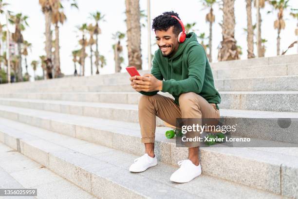 smiling young man using mobile phone while sitting on skateboard - jugendkultur sitzen konsole stock-fotos und bilder