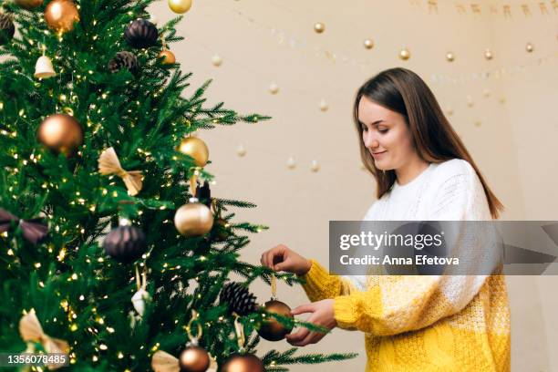 woman in white-yellow knitted sweater is decorating green christmas tree with bronze and gold baubles against white wall with garlands. new year celebration concept. front view - decorare l'albero di natale foto e immagini stock