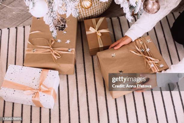 woman hands are holding gift box near white and beige paper gift boxes on carpet near decorated christmas tree in wicker basket. new year celebration concept. top view - anna kraft stock pictures, royalty-free photos & images