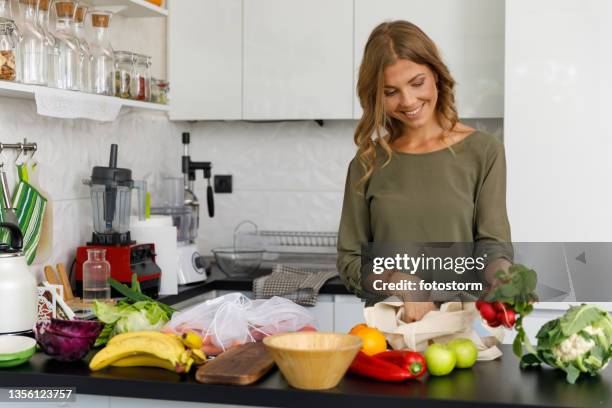 smiling woman unpacking fresh produce on her kitchen counter - unloading stockfoto's en -beelden