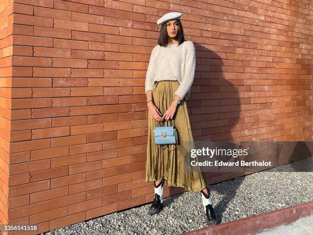 Model Jacqueline Barth wears a white felt / wool beret, a beige fluffy wool pullover, a silver watch, a high waist gold pleated / accordion midi...