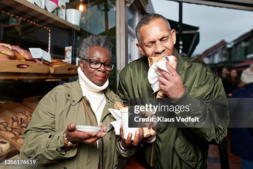 Senior man eating doughnut standing by female friend Senior man eating doughnut standing by female friend