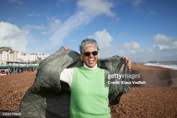 cheerful senior woman removing jacket at beach - green coat foto e immagini stock