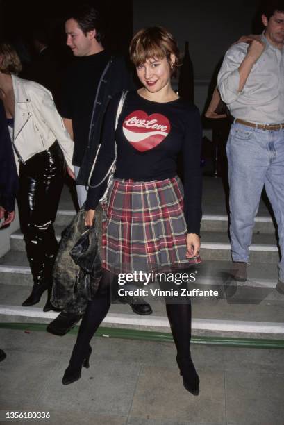 American-Canadian actress Jennifer Tilly, wearing a plaid skirt and a long-sleeved black top with an image parodying the 'Coca-Cola' logo on the...