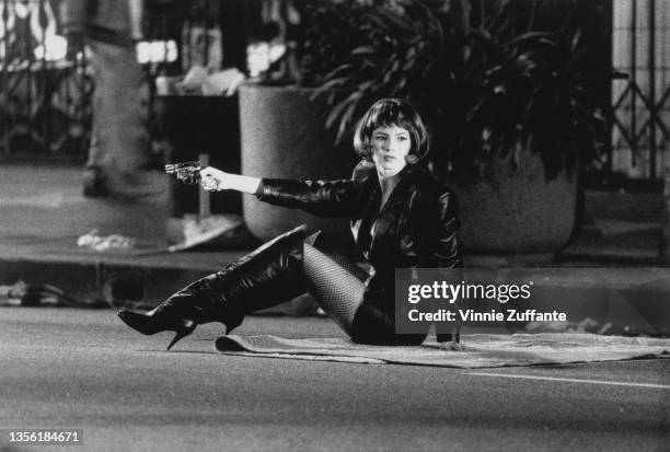 American actress Traci Lords, wearing knee-high boots, leather jacket and leather miniskirt, sits on the ground while aiming a revolver during the...