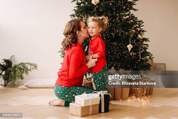 the concept of a happy family is preparing for christmas. cheerful mom and little daughter a child in red pajamas and red santa claus hats have fun hugging laughing sitting at a decorated christmas tree on new year's eve in a cozy indoor house - kerstboom versieren stockfoto's en -beelden