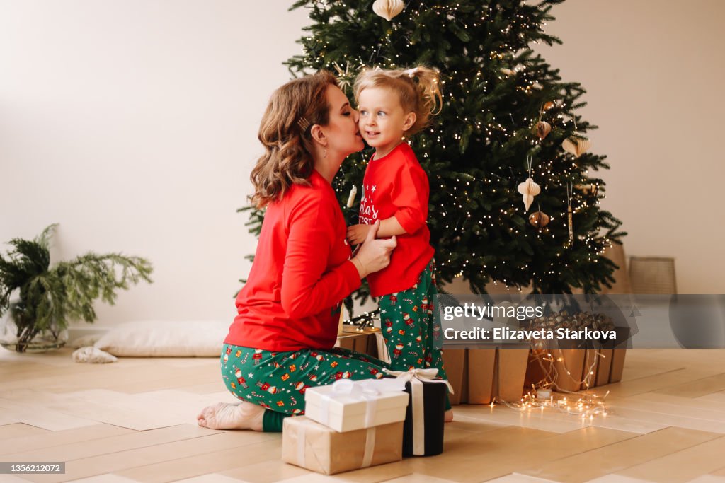 The concept of a happy family is preparing for Christmas. Cheerful mom and little daughter a child in red pajamas and red Santa Claus hats have fun hugging laughing sitting at a decorated Christmas tree on New Year's Eve in a cozy indoor house