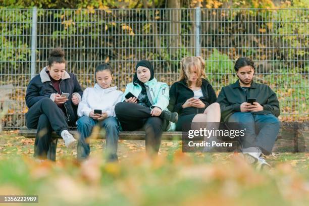 gruppe von teenager-freunden, die mit ihren mobiltelefonen auf der bank sitzen - ethnische integration deutschland stock-fotos und bilder