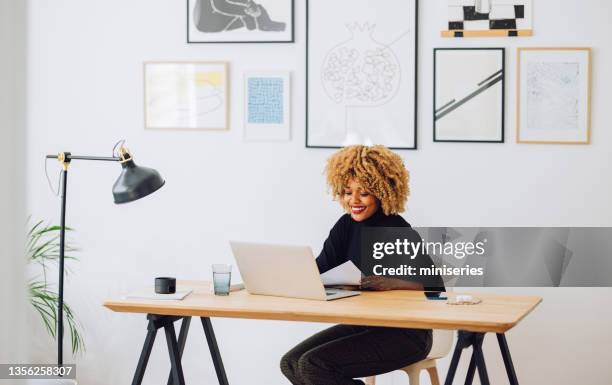 smiling afro-american woman sitting at desk in her office and looking at a laptop - wide angle people stock pictures, royalty-free photos & images