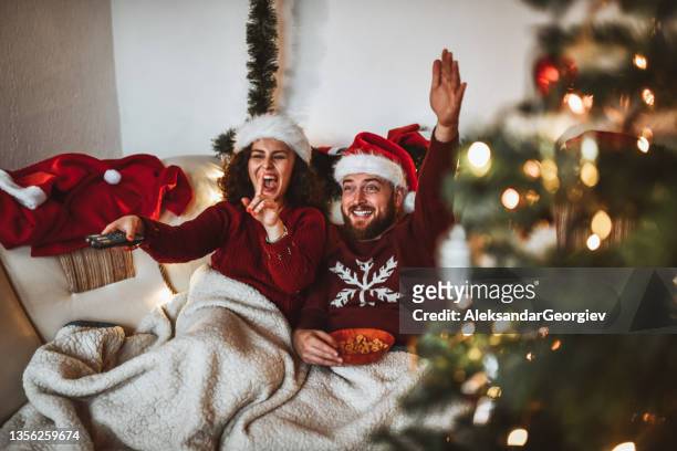 pareja sonriente con sombreros de papá noel celebrando la navidad comiendo bocadillos y viendo la televisión - lovers 2020 film fotografías e imágenes de stock