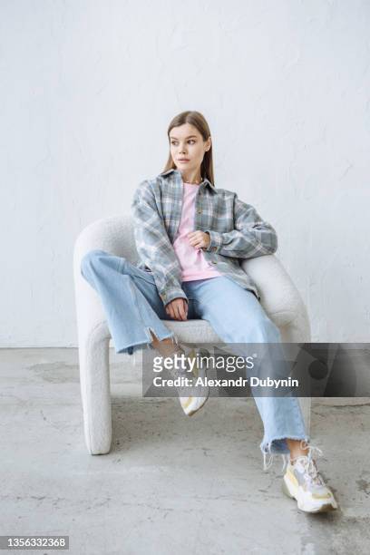 stylish young brunette woman posing sitting in a chair in casual clothes - fashion collection stock-fotos und bilder
