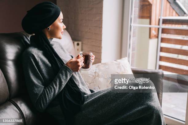 portrait of muslim woman in gray dress and black headscarf sitting on sofa with hot cup of tea and looking out of window - hot middle eastern women stock-fotos und bilder