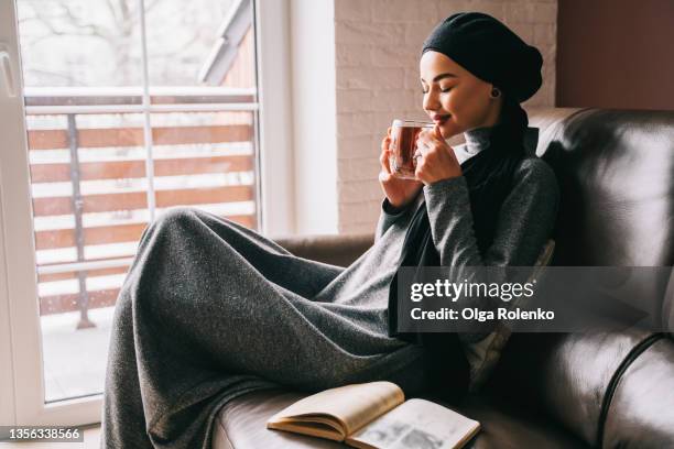 portrait of muslim woman in gray dress and black headscarf sitting on sofa with hot cup of tea and looking out of window - hot arab women stock-fotos und bilder