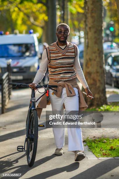 Guest wears sunglasses, a beige ribbed long sleeves pullover, a beige / brown / black striped sleeveless V-neck pullover, a beige wool coat, white...