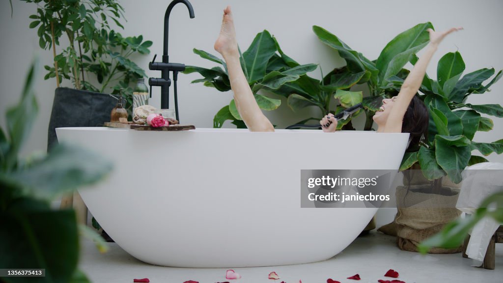 Young woman taking a bubble bath in lush green garden inside her bathroom. Singing to a hand shower, seductively washing leg