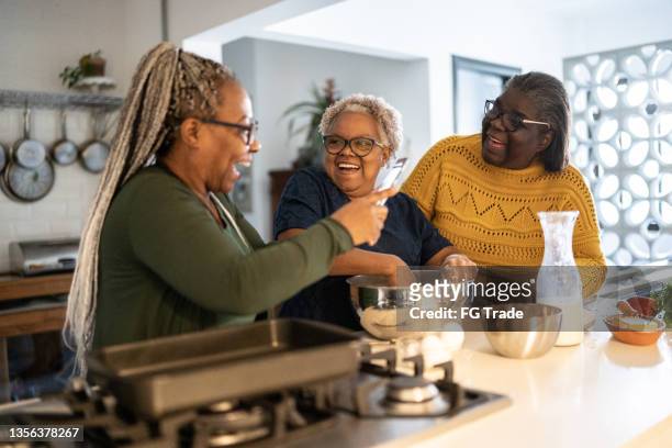 senior woman filming sisters or friends baking/cooking at home - 3 women senior kitchen stock pictures, royalty-free photos & images