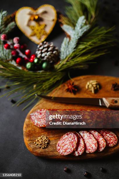 christmas low key flat lay of an homemade salami sausage served on a wooden cutting board with juniper berry, cardamom, clove, star anise, a kitchen knife and christmas decoration with an unfocused wooden heart and a golden glitter snowflake - delicatessen stock pictures, royalty-free photos & images