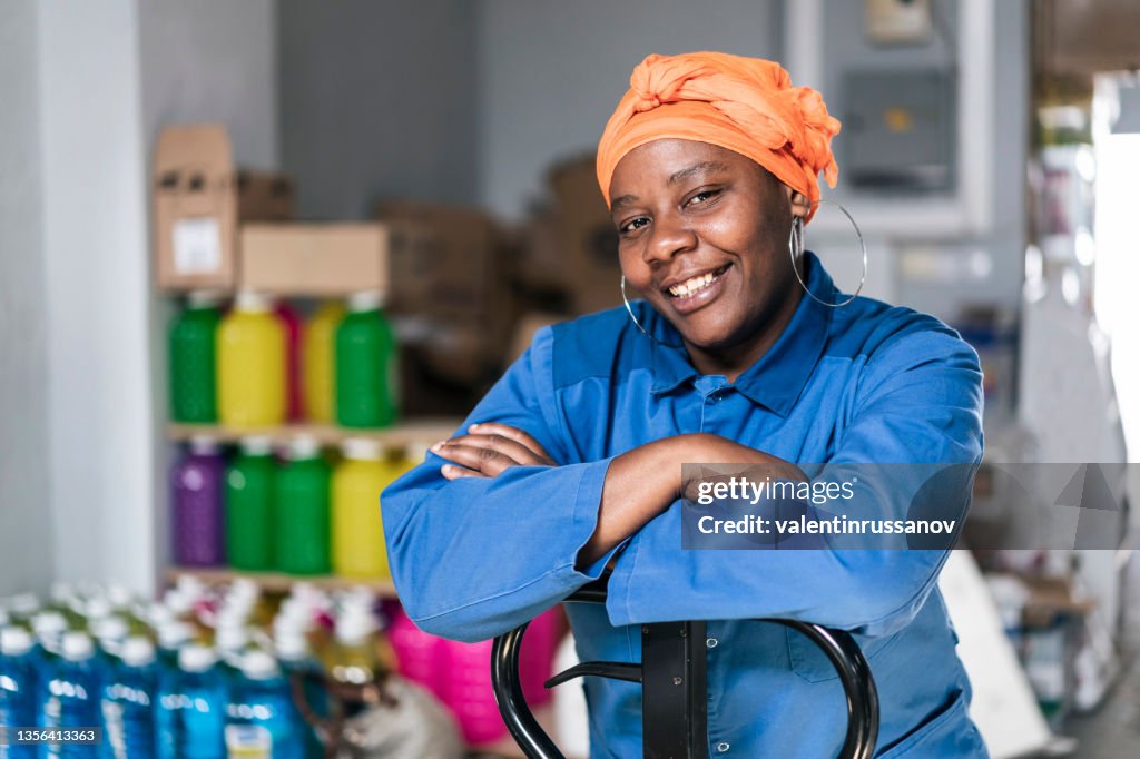 Mid adult african woman with hand pallet standing in a distribution warehouse. Portrait of a happy afro woman with headscarf, wearing uniform, working in a factory warehouse.