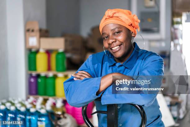 mid adult african woman with hand pallet standing in a distribution warehouse. portrait of a happy afro woman with headscarf, wearing uniform, working in a factory warehouse. - local market stock pictures, royalty-free photos & images