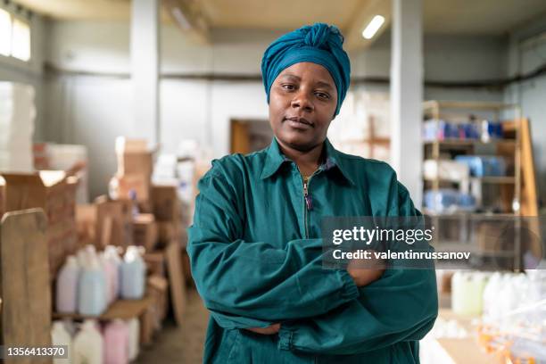 portrait of a confident afro woman with green head scarf and arms crossed, wearing working clothes on her work space - black culture stock pictures, royalty-free photos & images