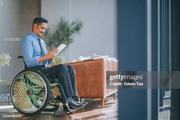 désactiver un col blanc indien asiatique en fauteuil roulant lecture tablette numérique souriant dans le salon - accessibilité aux personnes handicapées photos et images de collection