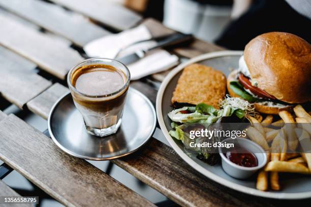 close up of a delicious cheeseburger, freshly served with fries and side salad on a wooden dining table, with a glass of coffee by the side in an outdoor restaurant - fastfood restaurant table stock-fotos und bilder