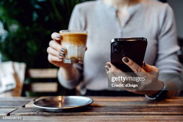 cropped shot, mid-section of young asian woman sitting at a table in coffee shop, looking at her smartphone while drinking a cup of coffee. lifestyle and technology - food bank bildbanksfoton och bilder