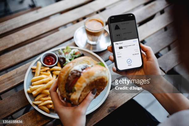 over the shoulder view of young asian woman shopping online on smartphone and making payment online with credit card while enjoying lunch in a restaurant. lifestyle and technology. comfortable and simple online shopping experience - meal expense stock pictures, royalty-free photos & images