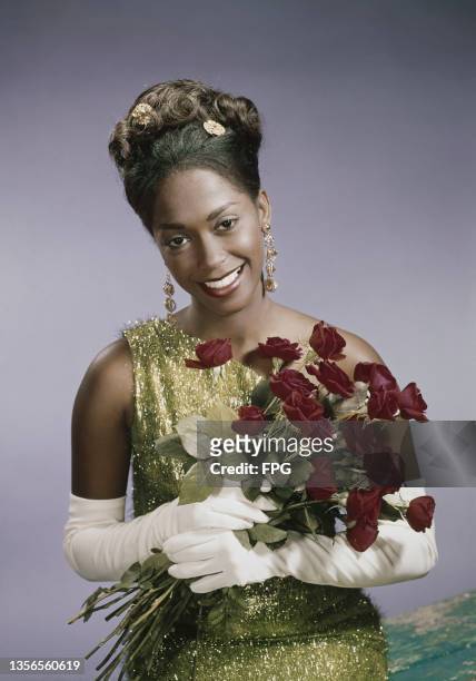 Woman wearing one-shoulder glitter gold dress and long white gloves holds a bouquet of red roses, studio shot, circa 1960.