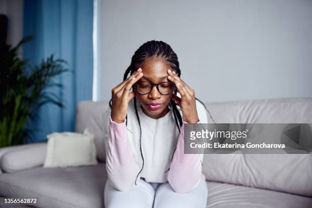 a young girl is sitting on the couch at home with her head in her hands. - angry black woman stock pictures, royalty-free photos & images