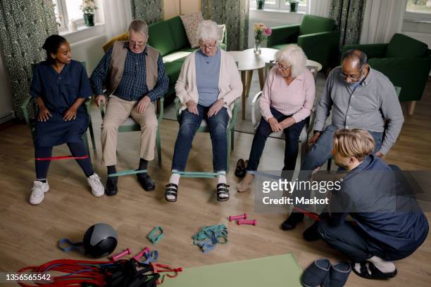 elderly people exercising with resistance band while male and female caregivers guiding them in living room - retirement community stock pictures, royalty-free photos & images
