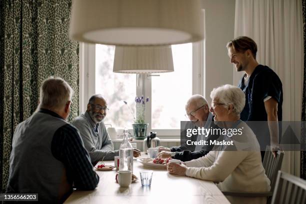 cheerful elderly woman and men talking with male caregiver having food at table - seniorenhuis stockfoto's en -beelden