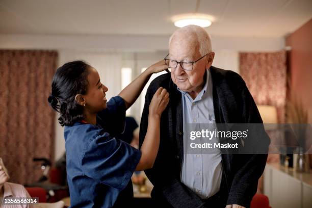 senior man looking at female nurse helping him to wear sweater in retirement home - retirement home stock-fotos und bilder