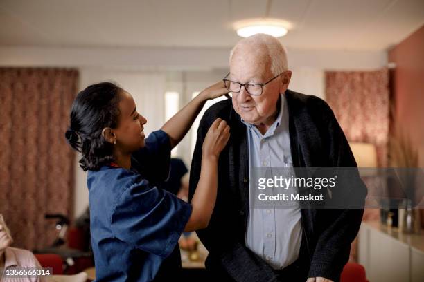senior man looking at female nurse helping him to wear sweater in retirement home - hospice photos et images de collection